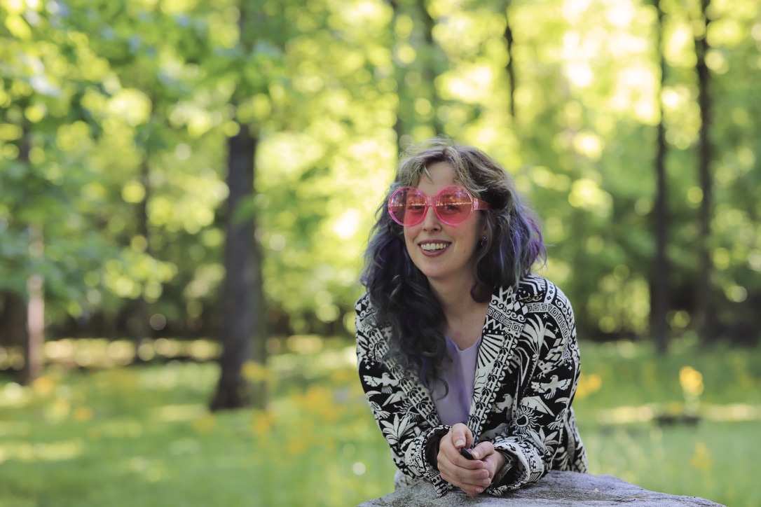 Aimee in pink sunglasses, smiling, in a sunlit forest